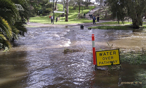 A photograph of a sign that reads : “Riverwalk Closed.” Clearly visible behind it is a flooded riverwalk.