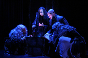 In a stage production of Macbeth, Macbeth, who is wearing a crown, gazes into the depths of a cauldron as the three witches tell him his future
