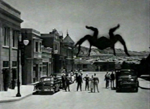 A movie still form the 1955 movie Tarantula! It shows the giant tarantula approaching the edge of town and people standing in the street looking at it.