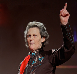 Photo of Temple Grandin. She wears a red short scarf and a western shirt. She is talking and pointing into the air with her index finger.