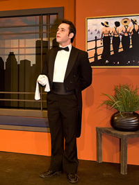 A photograph of a man playing a butler in a stage production. He is wearing a full tuxedo with a small towel draped over his forearm.