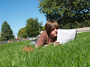 A photograph of a male student lying in the grass and reading a spiral bound text.