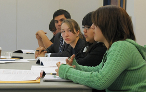 A photograph of a several young people sitting a table presenting or arguing a topic with documents and books spread out before them