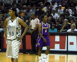 A photograph of two WNBA basketball players during a game. There are referees and a large seated crowd in the background