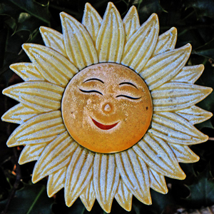 A photograph of a ceramic smiling sun. The sun could also be a sunflower. The face is the center of the sunflower.