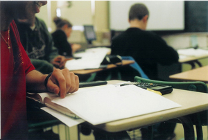 A photograph of a student sitting at a desk in a mathematics class. There is an open notebook and a calculator on the desk.