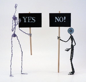 A photograph of two wire sculptures that represent humans. They are holding signs: one reads “Yes,” while the other reads “No!”