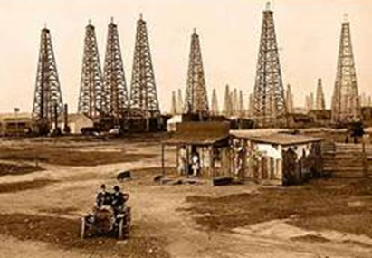 Image of an aged photo of an oil field lined with several oil derricks. A small shack is in the foreground, as well as a model-T ford with two occupants.