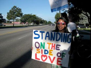 A young woman standing on the side of the road holding a sign that says 'Standing on the side of Love'