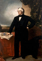 A man with parted grey hair standing with his right hand on a desk wearing a black suit.
