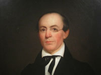 A portrait of a balding man with spectacles, wearing a black coat with a black string tie.