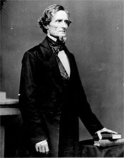 A photograph of a man standing with his left hand resting on a book wearing a black suit.