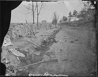 A photograph of a stonewall on the left and a ditch with many bodies in it and guns scattered