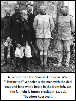 Five military officers in uniform standing in woods. On the far right is a young Theodore Roosevelt, a man in a white uniform with closely cropped hair and a mustache. In the front left is Joe Wheeler, a man with a white beard in a dark general’s coat, and white breeches.