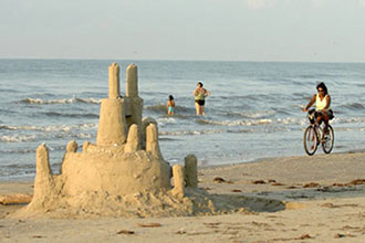Image of a beach with a sand castle in the foreground. There is a woman riding a bike on the beach and a mother and daughter standing in the gulf waters. 