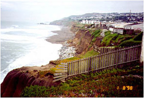 The first image is of a cliff area neighborhood in Pacifica, California. The neighborhood sits on a cliff overlooking the Pacific Ocean; behind the houses are fences and several yards of green that extend to the cliff. This photo was taken January 8, 1998. The second photo was taken February 2, 1998, after several storms. The yards of green between the cliff and the houses have diminished greatly. The houses are more exposed to the ocean.