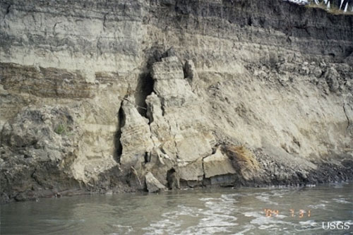 Photo of the bank of the Missouri River. The bank is made of rock and is erosion is evident. There are pieces of rock that are broken; there are ridges that indicated that water has reached various levels. Above the bank there is a tiny image of grass and trees.