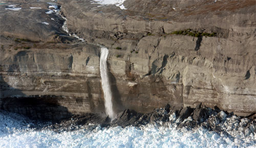Image of a cliff from the St. Elias mountain range in Alaska. There is snow above and below the cliff and a water fall made of the ice water coming from the top of the mountain falls down the side of the cliff to the mountains below. The cliff has evidence of erosion: the rock is various shades and the textures.