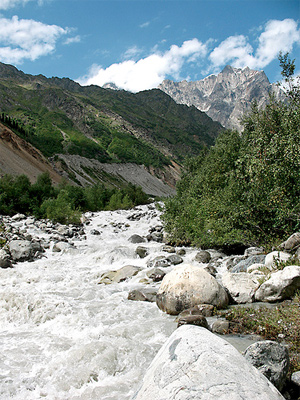 Image of a white water river that is flowing between a mountain range. There are trees and large shrubs at the base of the mountains.