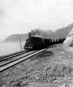 Image of a trainer on a rail line that runs alongside a body of water. There is thick smoke coming out of the chimney of the train.
