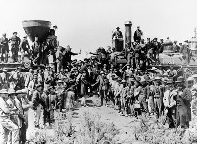 Image of two locomotives facing each other and at least one hundred men lined up one either side, facing the camera. Some men are standing or sitting on the locomotive. On the left locomotive a man hangs of the train holding a bottle; a man on the opposite holds a glass. There are two men on opposite sides shaking hands.