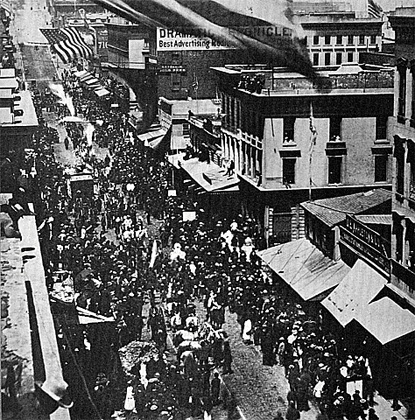 Image of crowds of people lined up along the two sides of a major street lined with tall buildings. An American flag is flying over one of the buildings. A parade of people and a few horse pulled carriages are in the middle.