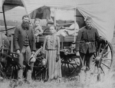 Image of a family (Adult male, adult female, adolescent male, and a toddler). There are standing in front of a covered wagon filled with various items.