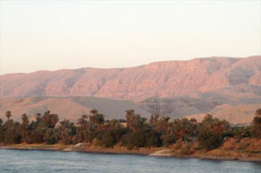 Image of the view along the Nile River at sunset south of Luxor. The land is green close to the river where is can be irrigated. The background is desert and mountain.