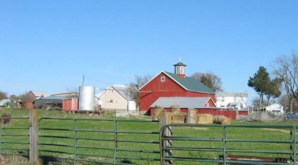 Image of a farm. There is a barn and several bails of hay. There is alos a silo.