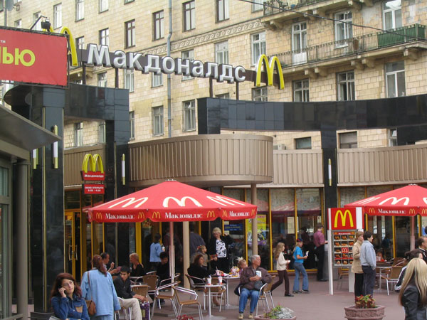 Image of a Russian McDonald's restaurant with sidewalk seating and umbrellas with the golden arches and McDonald's written in Russian. 