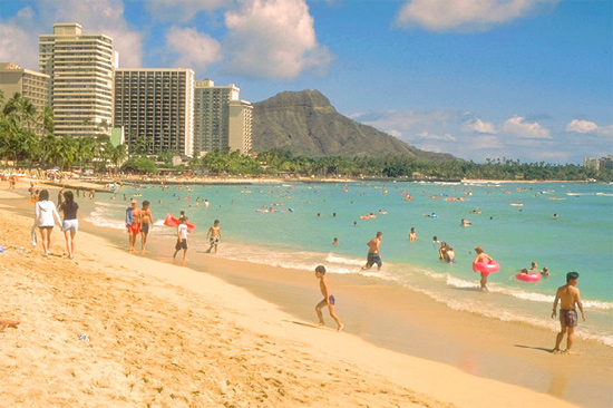 Image of a scene of a Hawaiian Beach. People are walking along the beach and some are swimming in the water. There are a row of  hotels in the background and a mountain can be seen beyond them.
