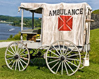 Image of a white, covered carriage with four wheels (it would have been pulled by a horse during the Civil War. It is labeled 'Ambulance' and a red cross is below.  A wooden gurney sticks out of the back.