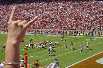 Image of a person’s hand displaying the Hook 'Em Horns' sign. A football game and the crowded stadium are in the background. 