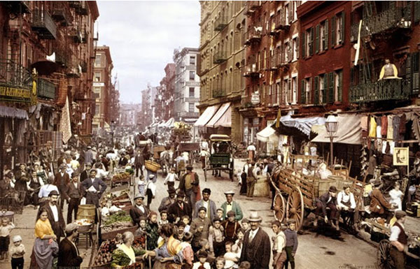 Image of Mulberry Street taken in the early 1900s. The street is crowded on both sides with various people and activities. The streets are lined with fresh produce vendors. The sidewalks are also crowded with people walking, but also with shops and vendors on the sidewalk. There are also horse-drawn carriages on the street.