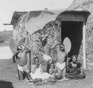 Image of native Hawaiians eating poi in front of a grass hut. There are two men kneeling and one adolescent male dressed in only a loin-covering. There is one child seated; there is an adolescent female and adult female seated.