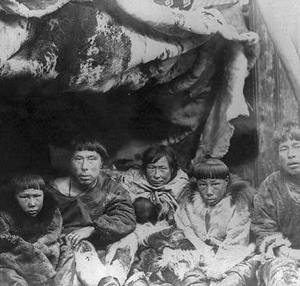 Image of a family of Alaskan Natives sitting in a tent covered in animal skins. There are two children who are each dressed in fur coverings. There are three adults (two men and one woman) who are draped in leather and fur.
