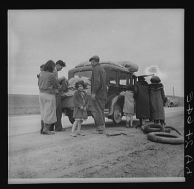 Image of a family of seven around a car that is missing a tire on the back passenger side.