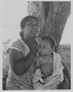 Image of a Mexican mother sitting under a tree, holding a baby who is draped in a blanket.
