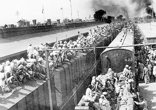 Image of a rail yard with two trains; many people are sitting on top of the cars of the train on the left. There are people lined up to enter the train on the right, and several rows of people are sitting on the flatbed on the right train.