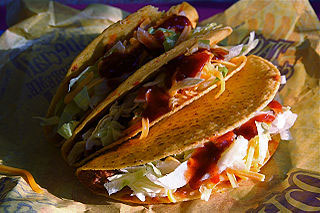 Image of three Taco Bell tacos; the tacos are crispy tortillas, with ground beef, lettuce, and cheese and topped with a sauce. The tacos are laid on a taco wrapper.