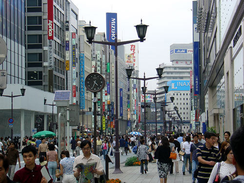 Image of a shopping district in downtown Tokyo. There are many people walking in the shopping district that is surrounded by tall buildings.