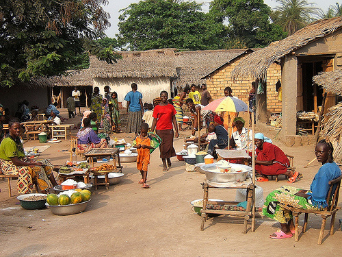 Image of a village marketplace surrounded by structures made of mud with straw coverings; there are several traders seated with their goods for sale while other people are walking around the market.