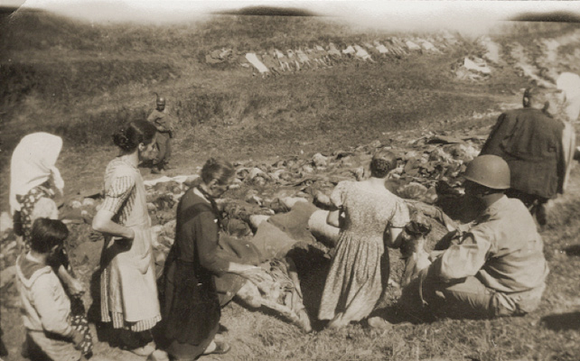 German civilians from Nammering are forced by U. S. troops to look at the corpses exhumed from a mass grave near the town: In the background there are bodies lined across the hillside. In the foreground, an American soldier sits as men, women, and children are paraded in front of a mass grave