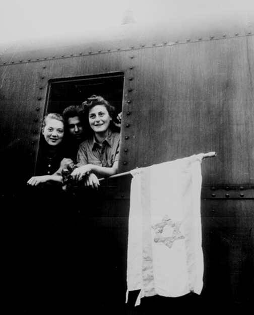 Image of two teenaged girls and a teenaged boy peeking out of the window of a ship. One of the girls is holding a tattered Jewish flag.
