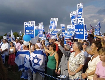 Image of men and women standing behind a banner of an Israeli flag. Many people in the crowd are holding signs that read 'Yes to Peace; No to terror' Others are holding blue and white balloons.