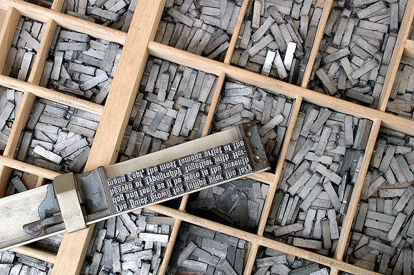 A photograph of movable type letters placed in a block ready for printing. In the background are boxes containing various movable type letters.