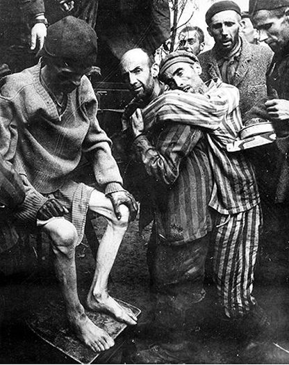 A group of prisoners inside a Nazi death camp. They are all very thin and wearing stripped pajama like uniforms. One of the men is carrying another who is obviously too weak to walk.