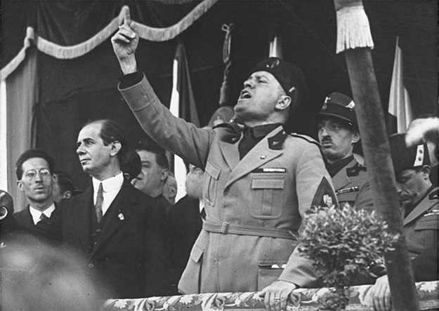 A photograph of Benito Mussolini at a rally speaking with his right arm raised for effect.