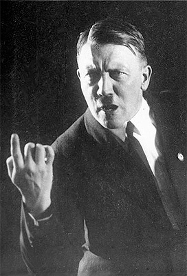 A photograph of Adolf Hitler speaking. He is wearing a suit and tie and has his right arm and index finger extended for effect.