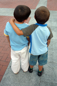 A photo of two small boys' backs as they stand and observe something out of the camera's view; One boy has his arm around the other one.
